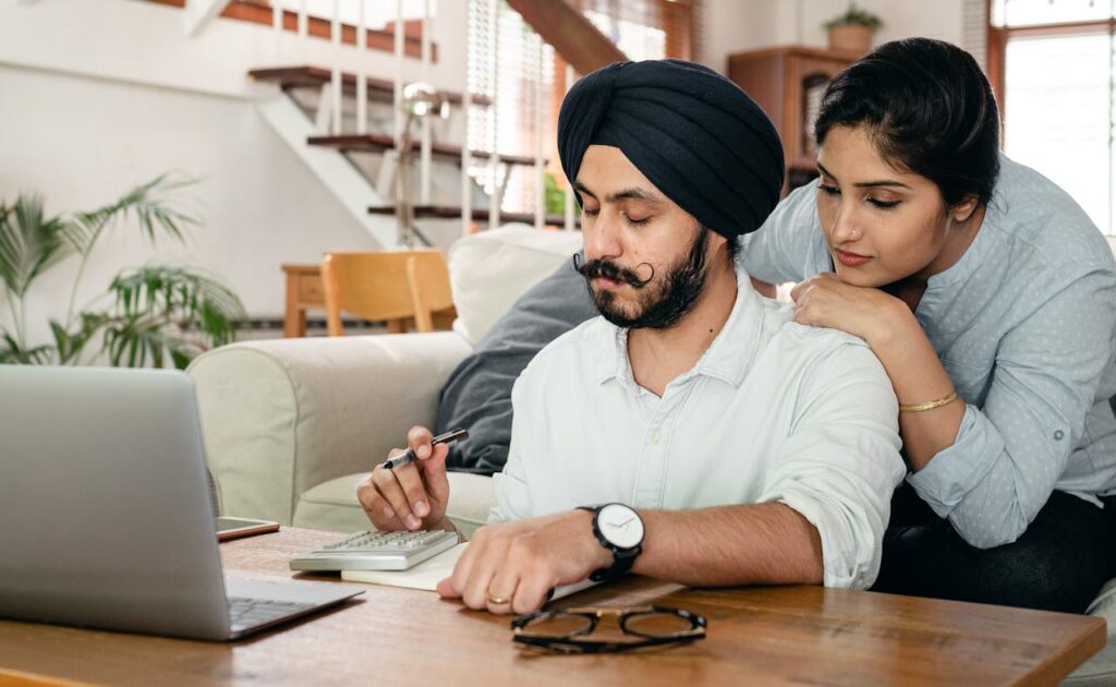 Focused Indian woman standing behind man in turban at table counting with calculator while writing notes in notepad